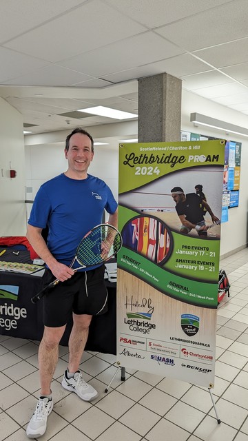 Daniel with a squash racquet in front of a Lethbridge 2024 squash tournament poster