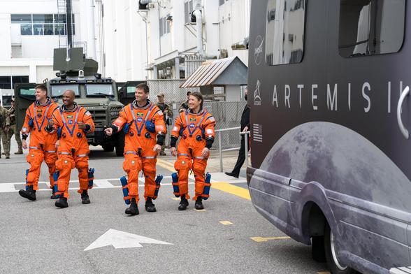 Vier Astronauten in orangefarbenen Anzügen, daneben ein Transporter mit der Aufschrift Artemis II